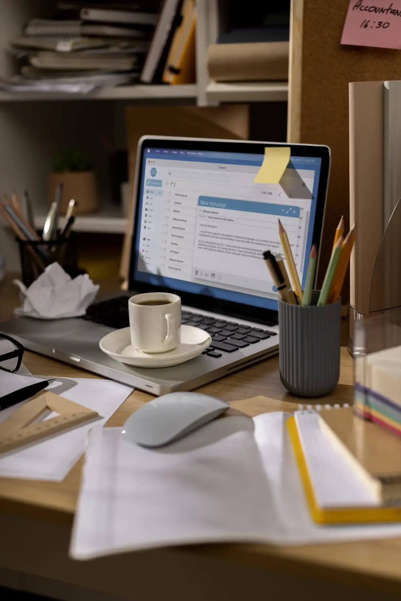 A cluttered office desk with an open laptop showing email, a coffee cup, scattered papers, a mouse, and pencils; the warm scene feels busy and focused