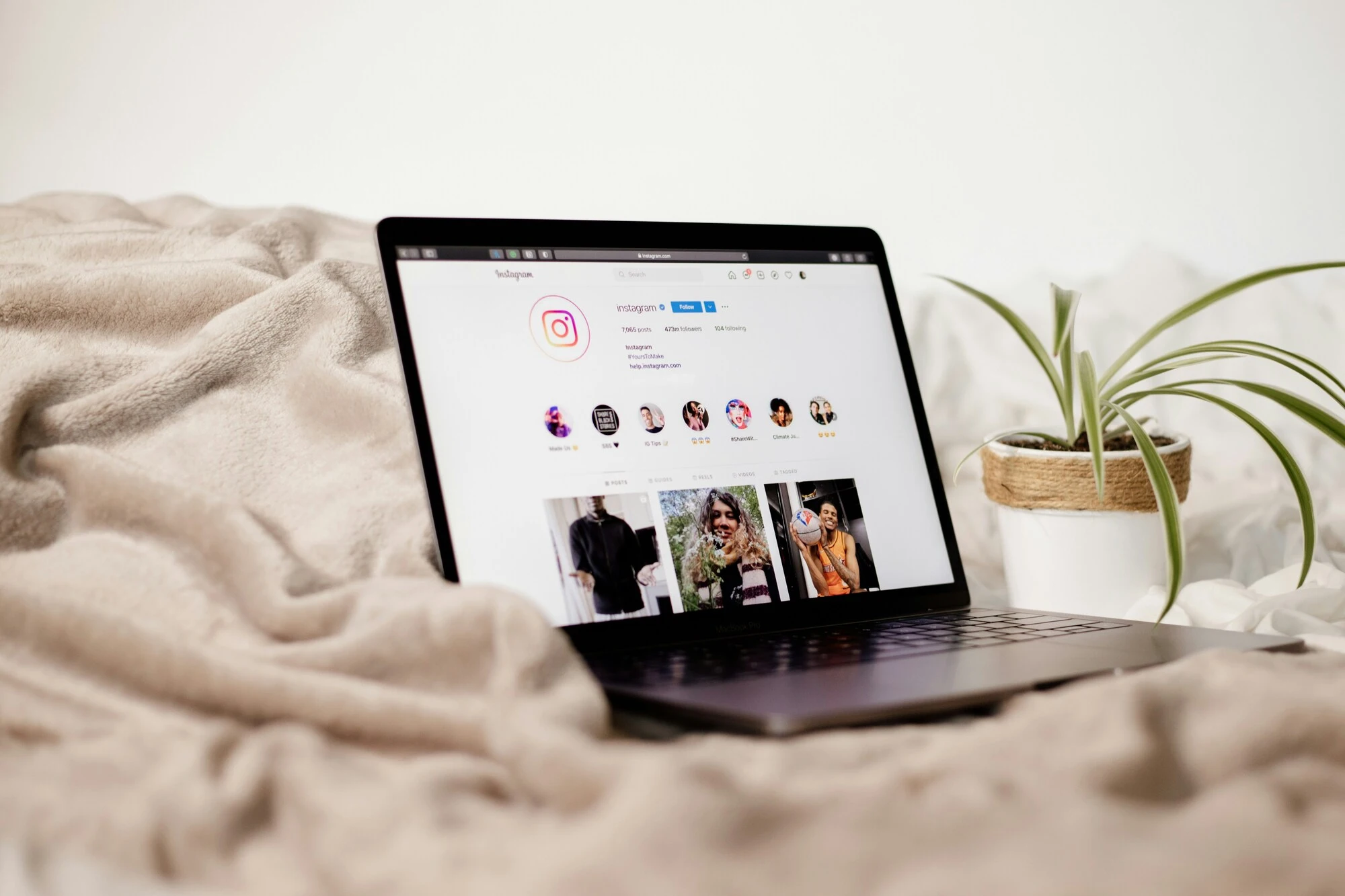 A laptop displaying an Instagram profile sits on a cozy beige blanket next to a potted plant, creating a relaxed, comfortable atmosphere