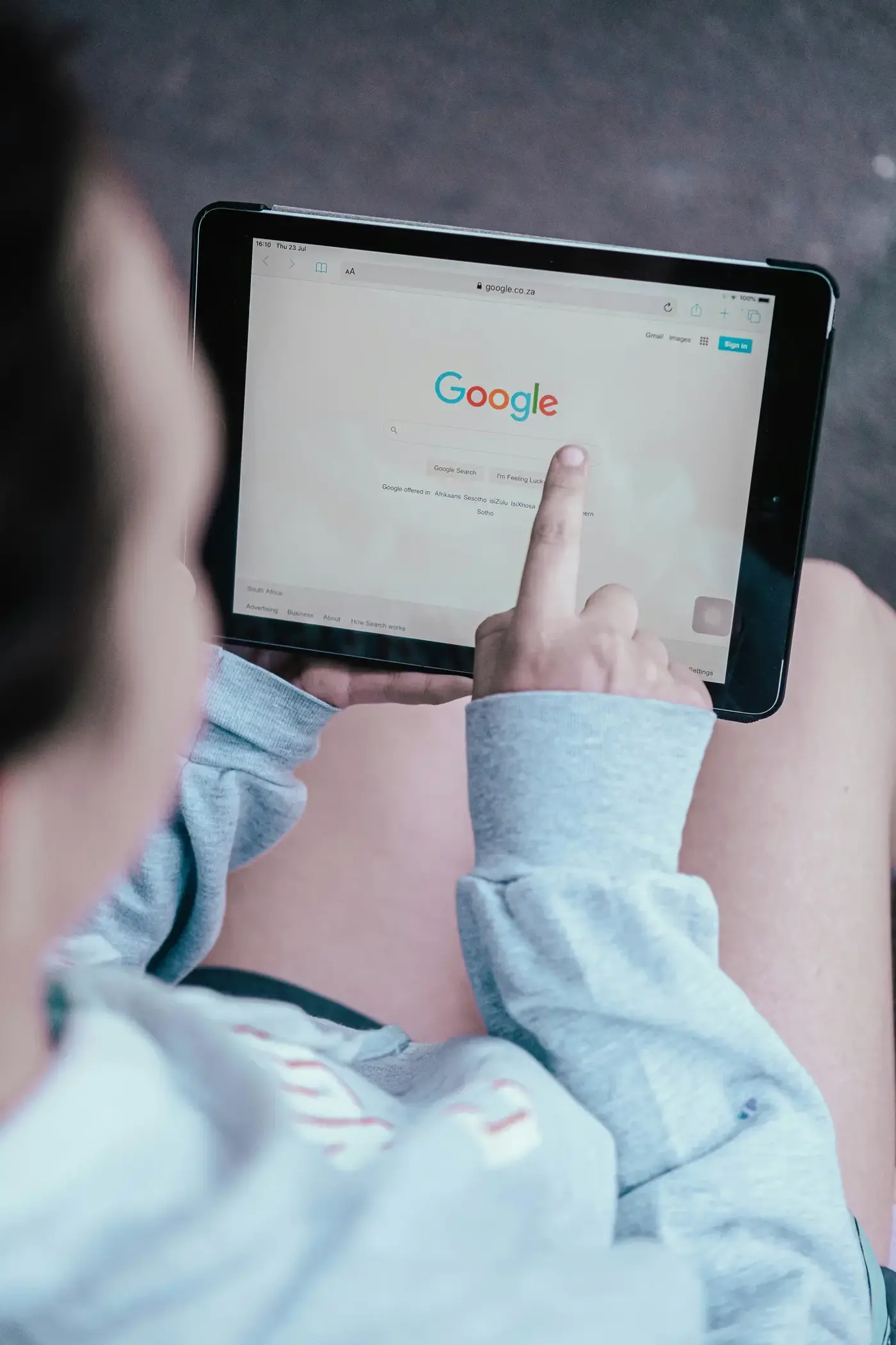 A person in a gray sweatshirt taps the Google search page on a tablet resting on their lap, suggesting casual browsing in a quiet setting