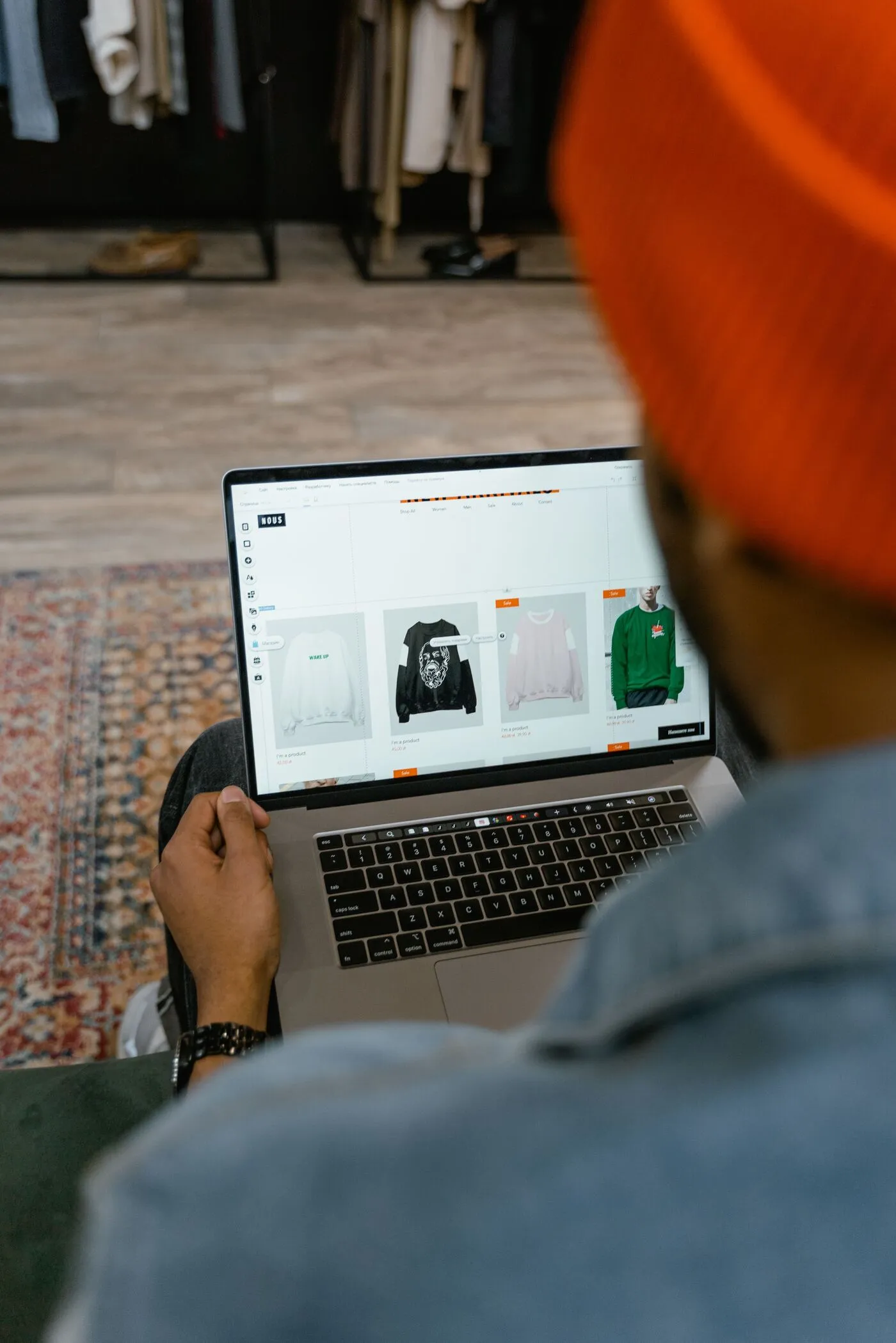 A shopper in an orange beanie browses clothing listings on a laptop in a clothing store, comparing online styles in a relaxed, casual setting