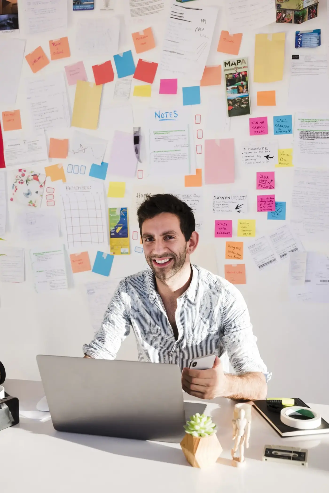 A smiling man works at a laptop beneath a wall covered with notes, sketches, and colorful sticky tabs, creating a busy, creative office atmosphere