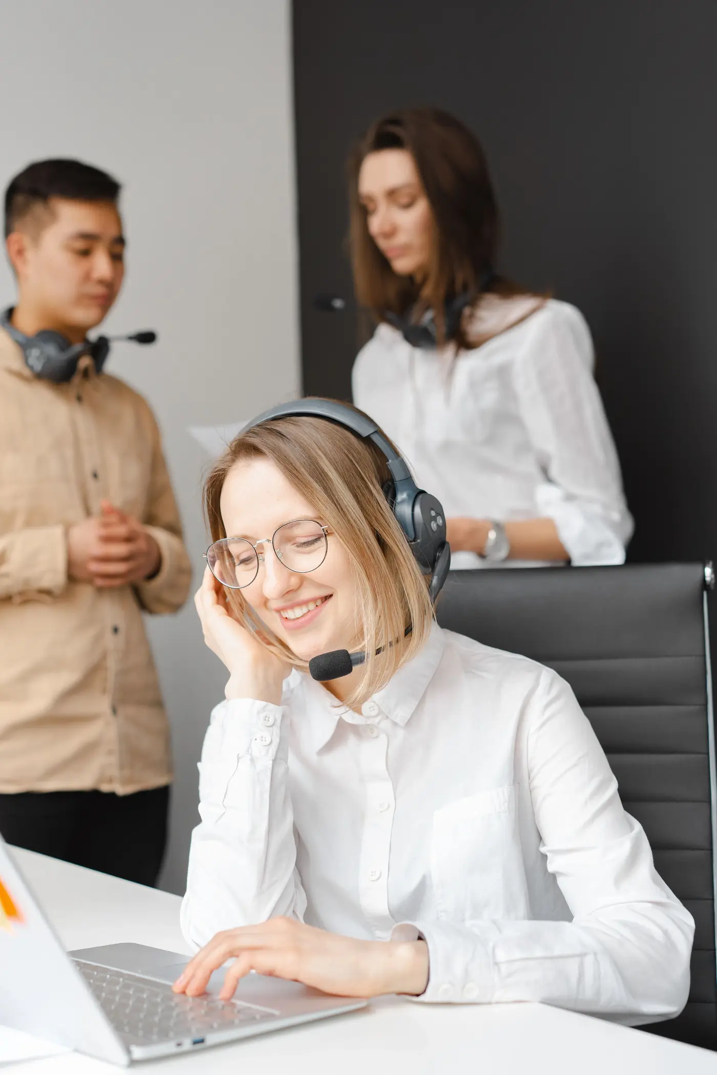 A smiling woman wearing a headset works at a laptop in a bright office, while two coworkers stand behind her, suggesting a friendly customer support team