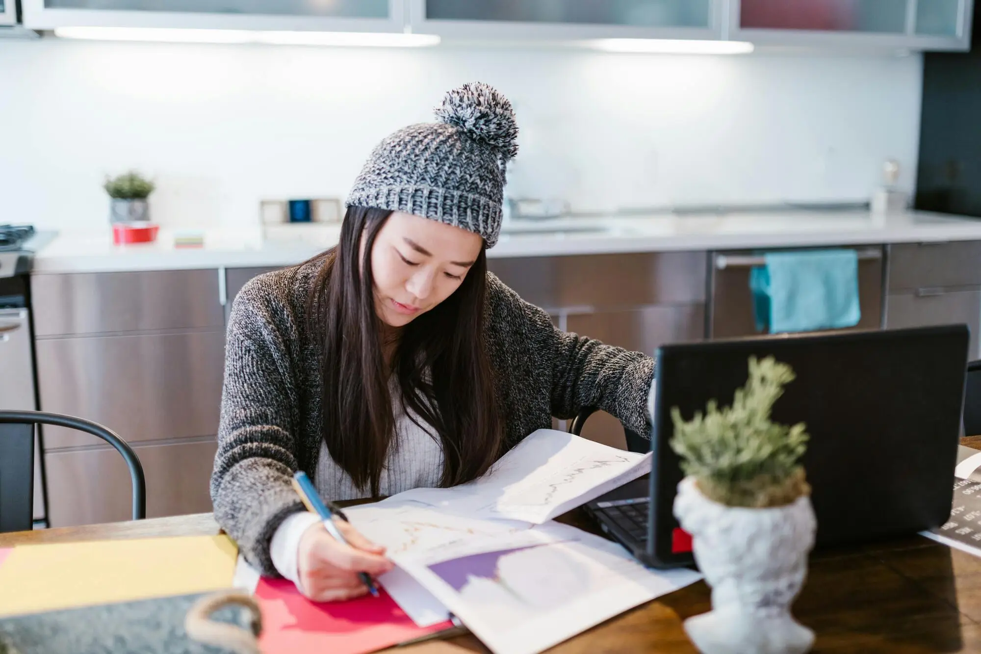 A woman in a knitted hat studies at a table cluttered with papers and a laptop in a modern kitchen, conveying focus and productivity