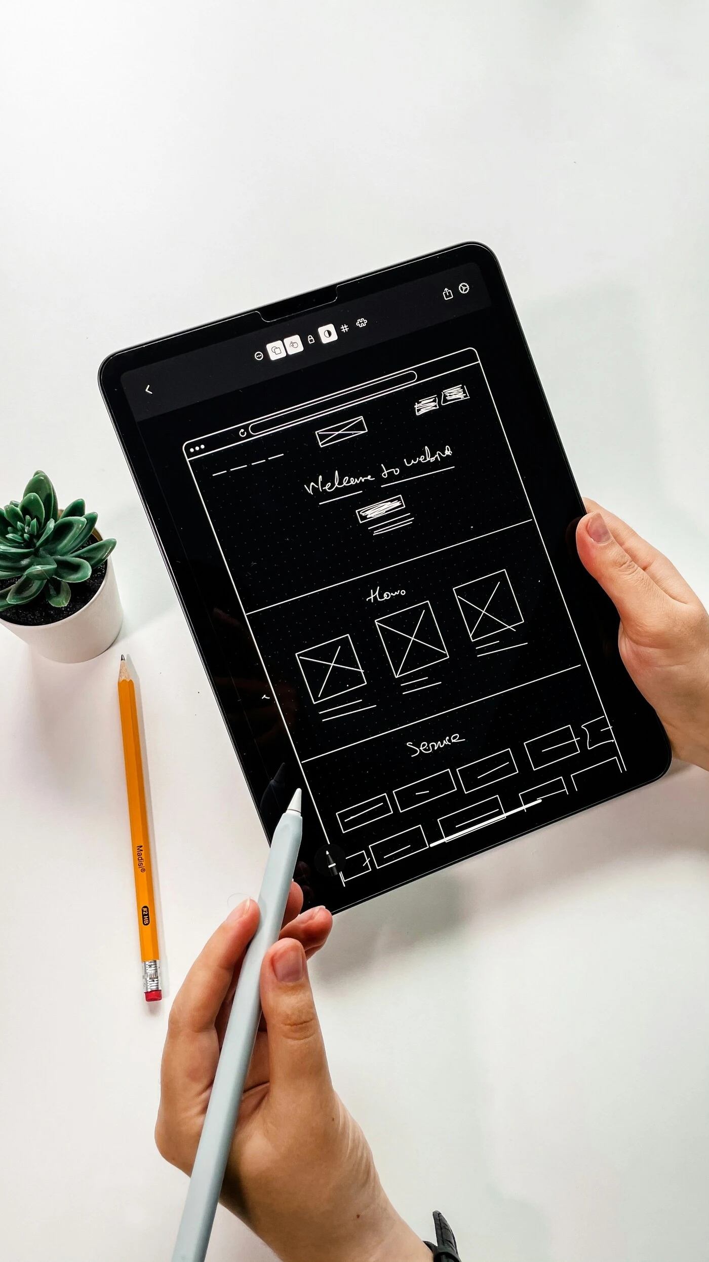 Overhead view of hands sketching a wireframe on a tablet with a stylus; a succulent and orange pencil sit nearby, creating a calm, focused workspace