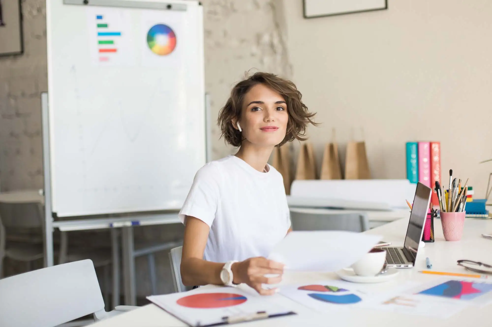 Young woman in a white shirt sits at a desk with colorful charts. A whiteboard with graphs is behind her. She looks confident and focused