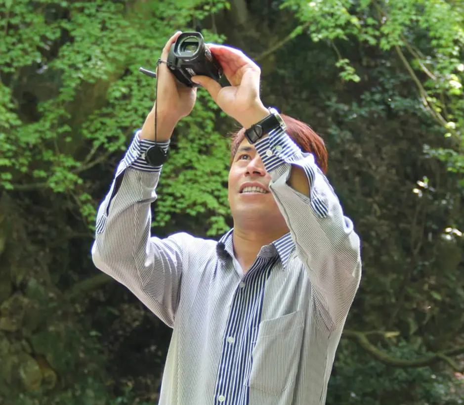 A man in a striped shirt, holding a camera upwards with a smile, stands in a sunlit forest