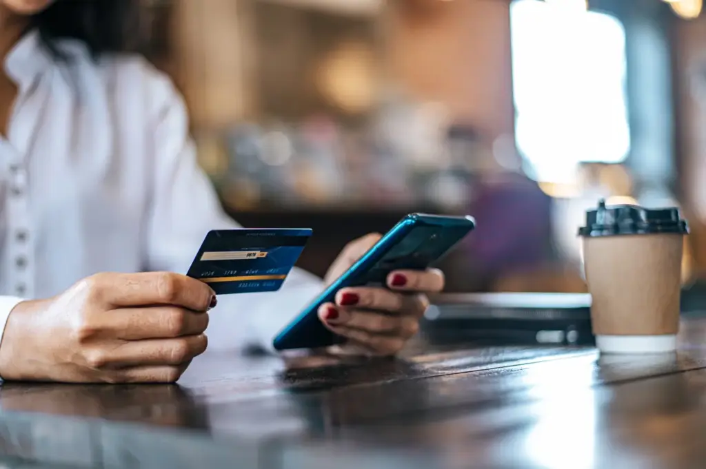 Person at a café holds a credit card and smartphone beside takeaway coffee, suggesting contactless payment, shopping, or online ordering