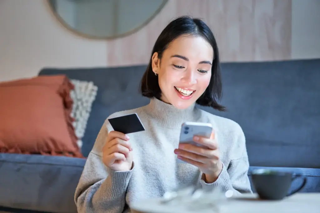 Smiling woman in a cozy living room holds a credit card and checks her smartphone, suggesting online shopping or a digital payment