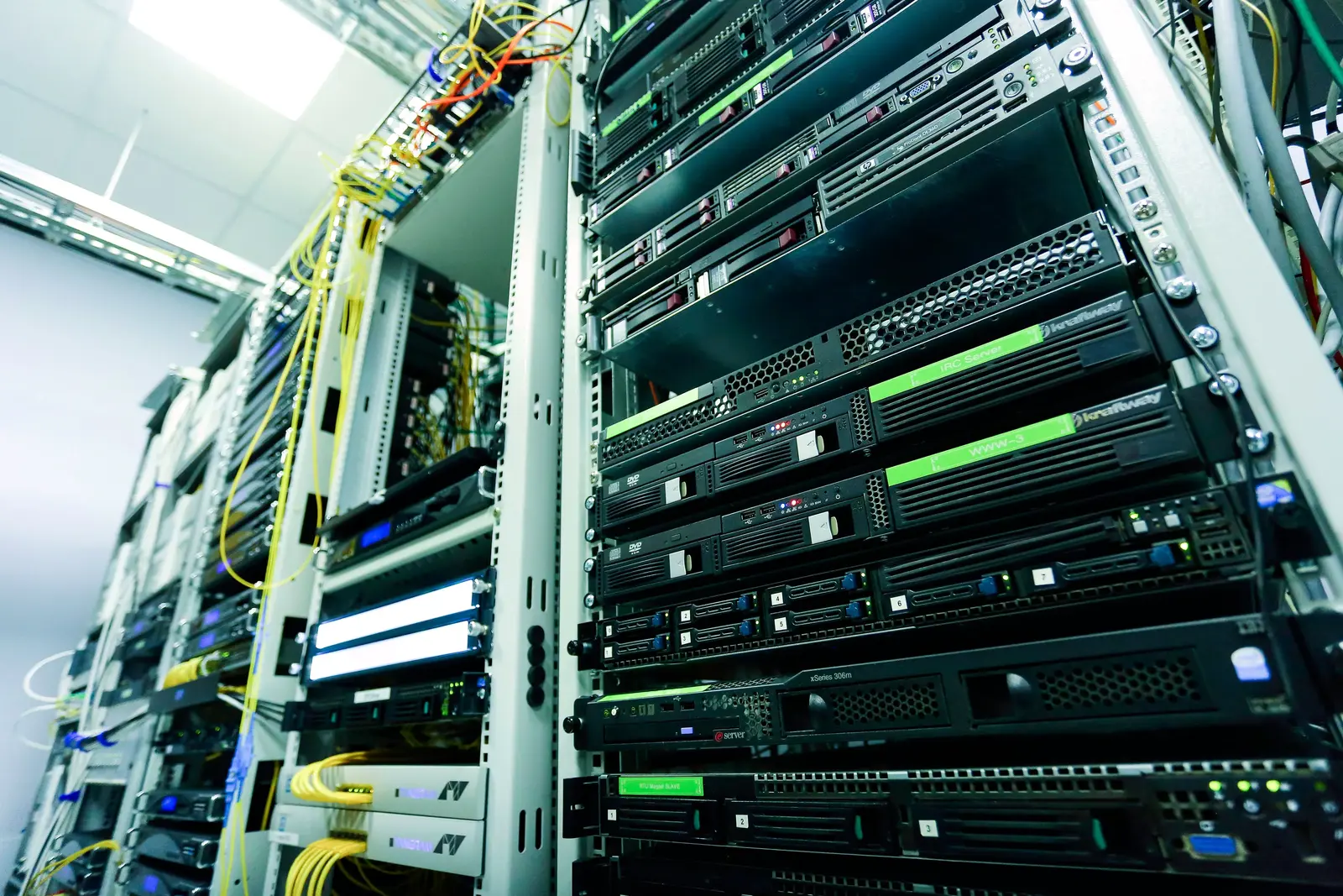 Tall server racks packed with black hardware, glowing green status lights, and tangled yellow cables in a bright data center, conveying a high-tech, busy atmosphere
