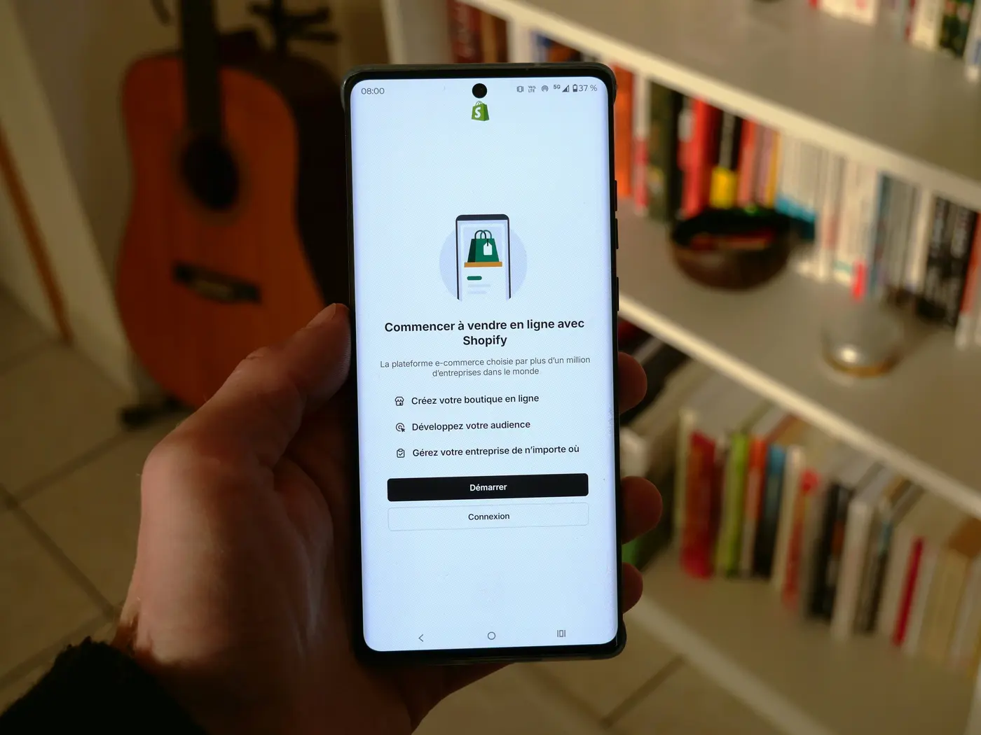 A hand holds a smartphone showing Shopify’s French online-store setup screen, with a guitar and colorful books on shelves in a warm room