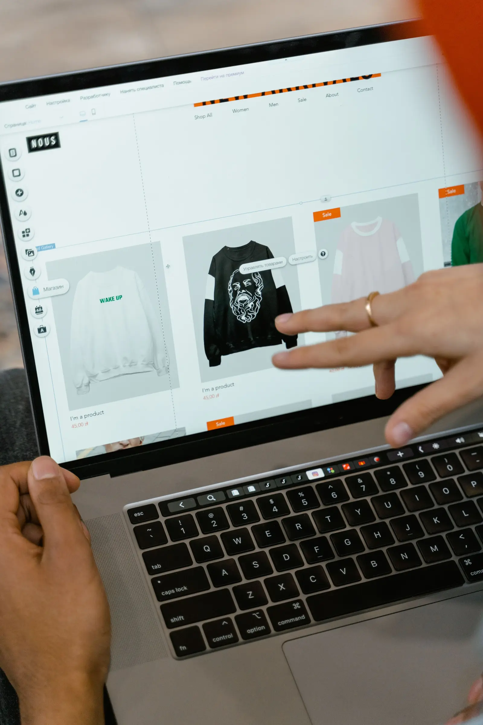 A person points to a black graphic T-shirt on a laptop screen displaying an online clothing store, with other apparel listings nearby