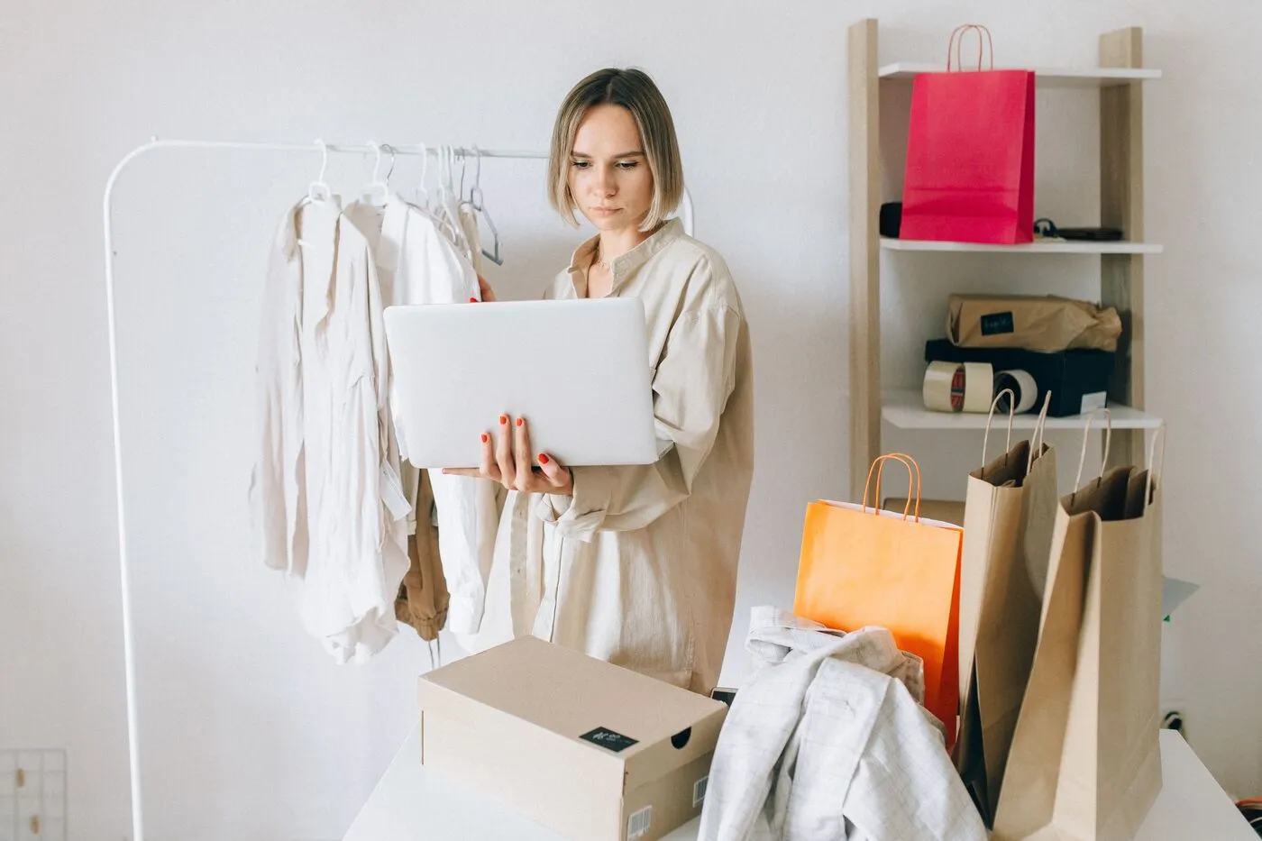 A woman reviews a laptop beside clothing racks, an open shipping box, and shopping bags in a bright room, suggesting online fashion shopping or returns