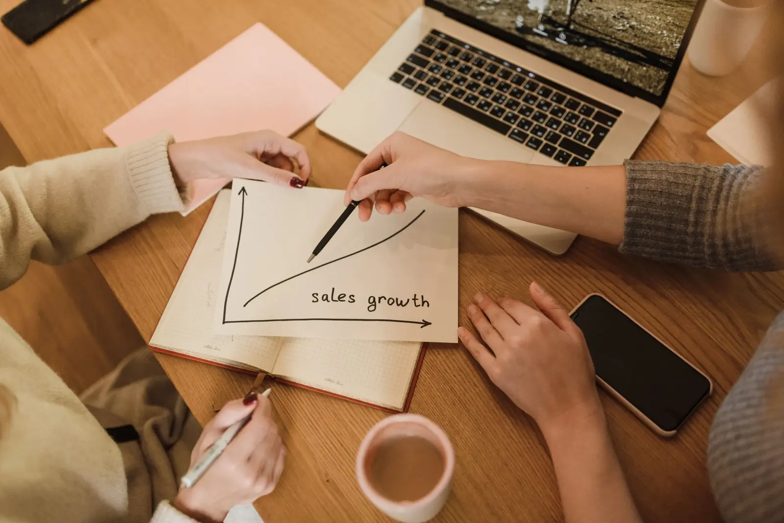 Coworkers discuss a hand-drawn sales growth chart in an open notebook, pointing at the upward trend beside a laptop and coffee on a wooden table