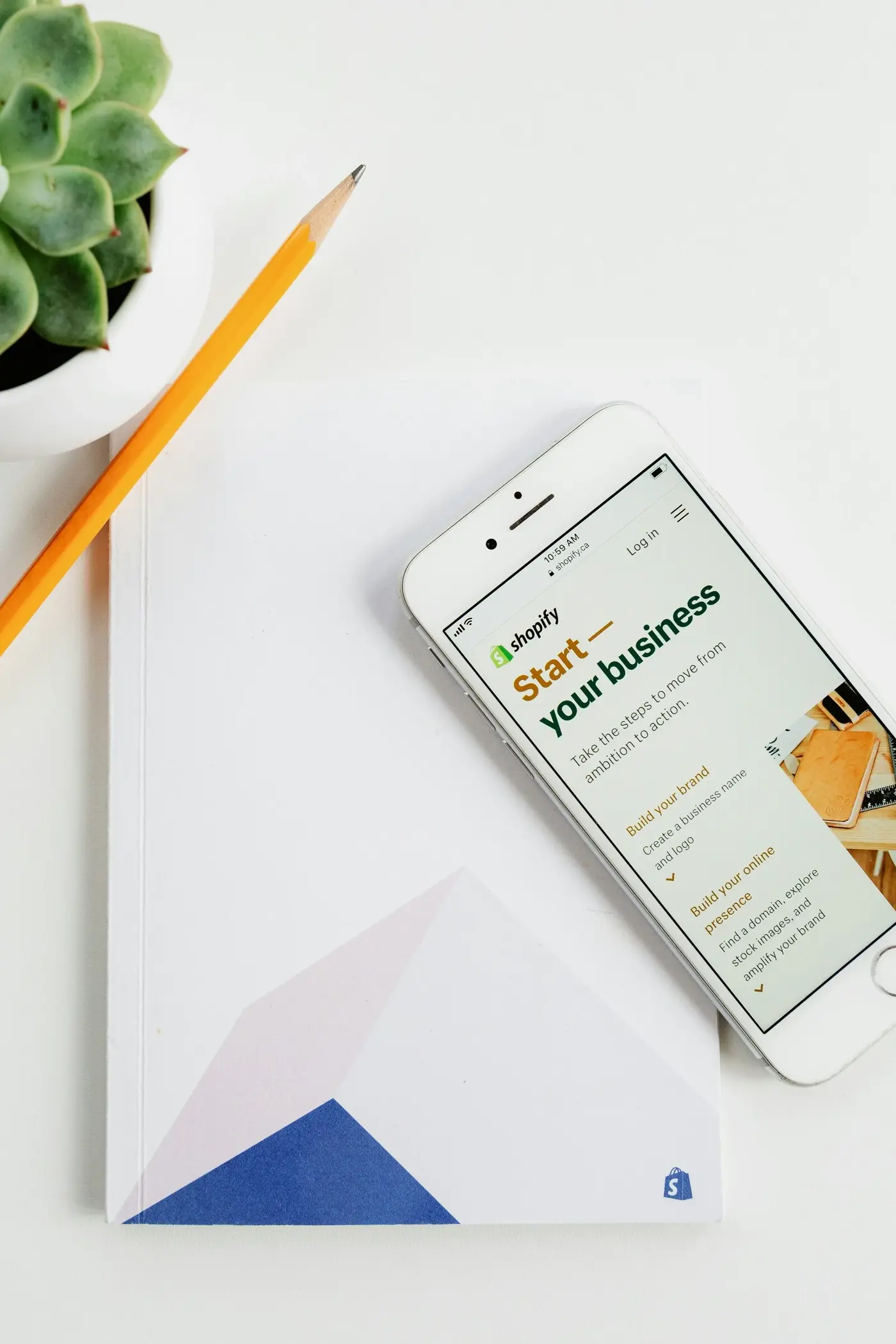 Minimalist workspace with a smartphone displaying a Shopify “Start your business” page beside a geometric notebook, yellow pencil, and succulent; bright, calm, and inspiring